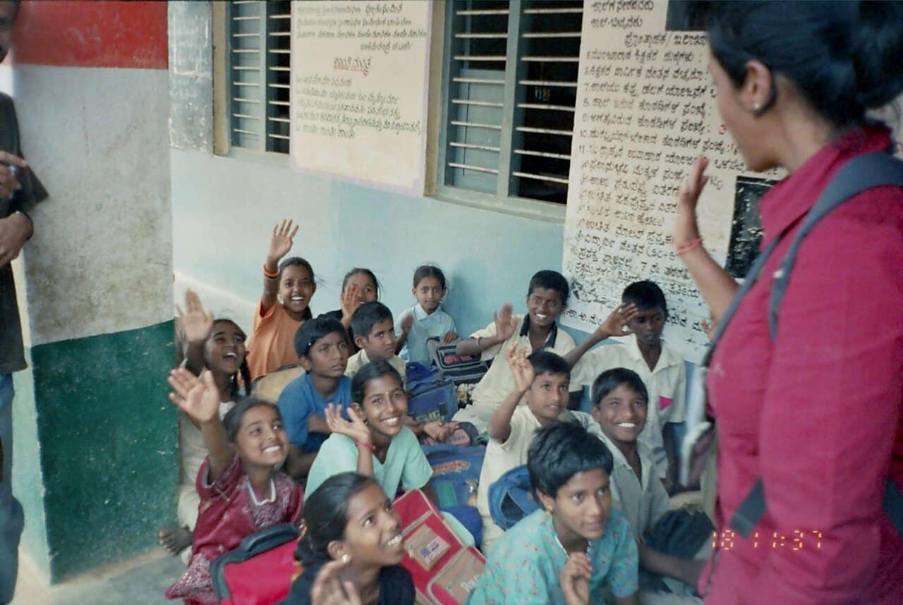 Schoolchildren seated on the floor of a classroom, raising their hands.