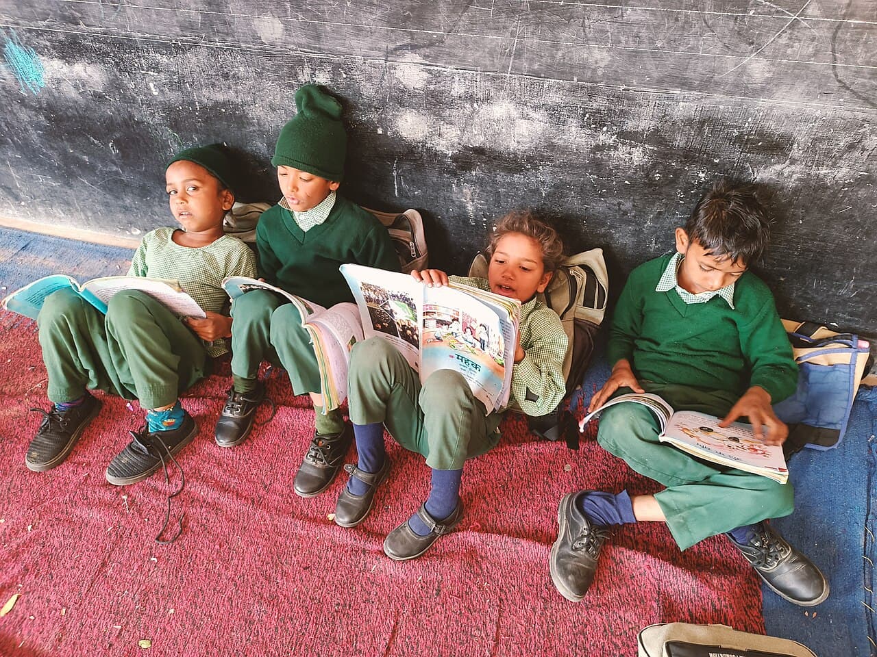 Four students in green uniforms reading books while seated against a blackboard.